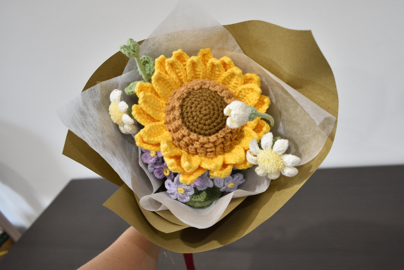 Handmade crochet flower bouquet featuring a single sunflower, with purple and white accent flowers and greenery, wrapped in brown paper and held against a neutral background.