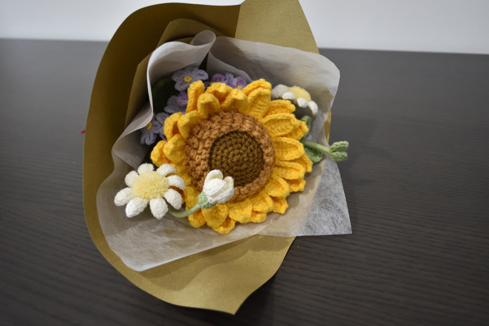 Handmade crochet flower bouquet featuring a single sunflower, with purple and white accent flowers and greenery, wrapped in brown paper and laid on a dark table.