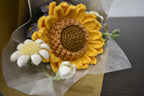 Close-up of a handmade crochet flower bouquet featuring a single sunflower, with purple and white accent flowers and greenery, wrapped in brown paper.