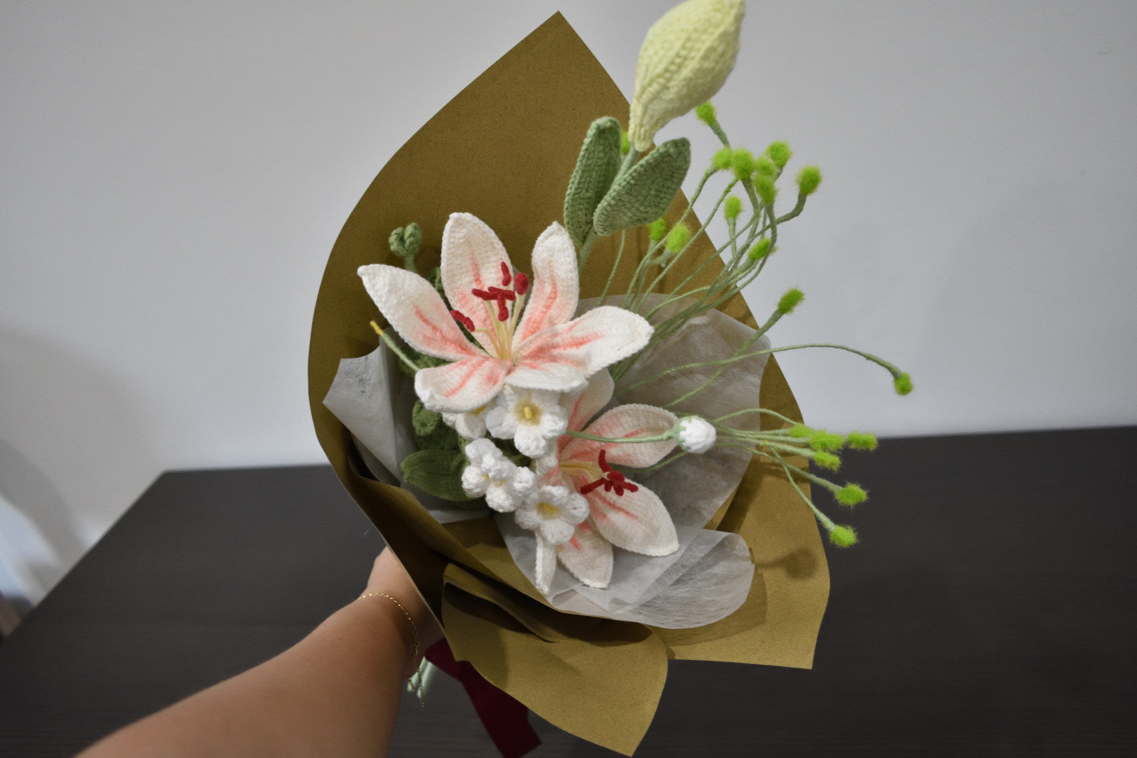 Handmade crochet flower bouquet featuring twin pink and white lilies with one flower bud, wrapped in brown paper and held against a neutral background.